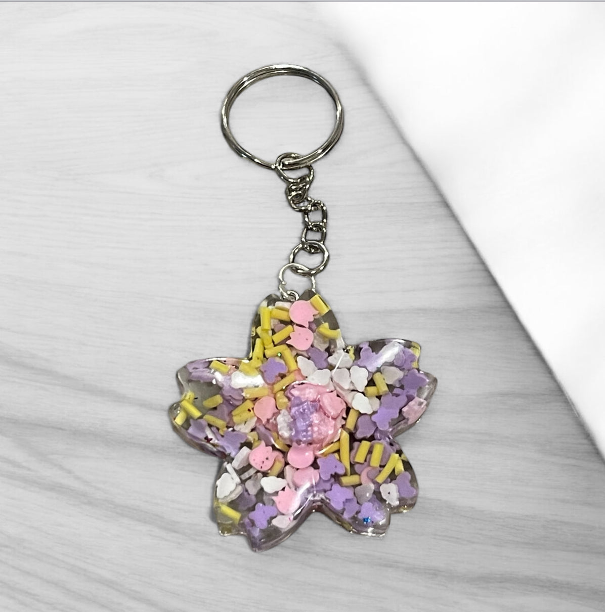 Handmade resin flower keychain with pastel purple, pink, and yellow sprinkles, featuring a delicate sakura blossom design with silver-tone hardware