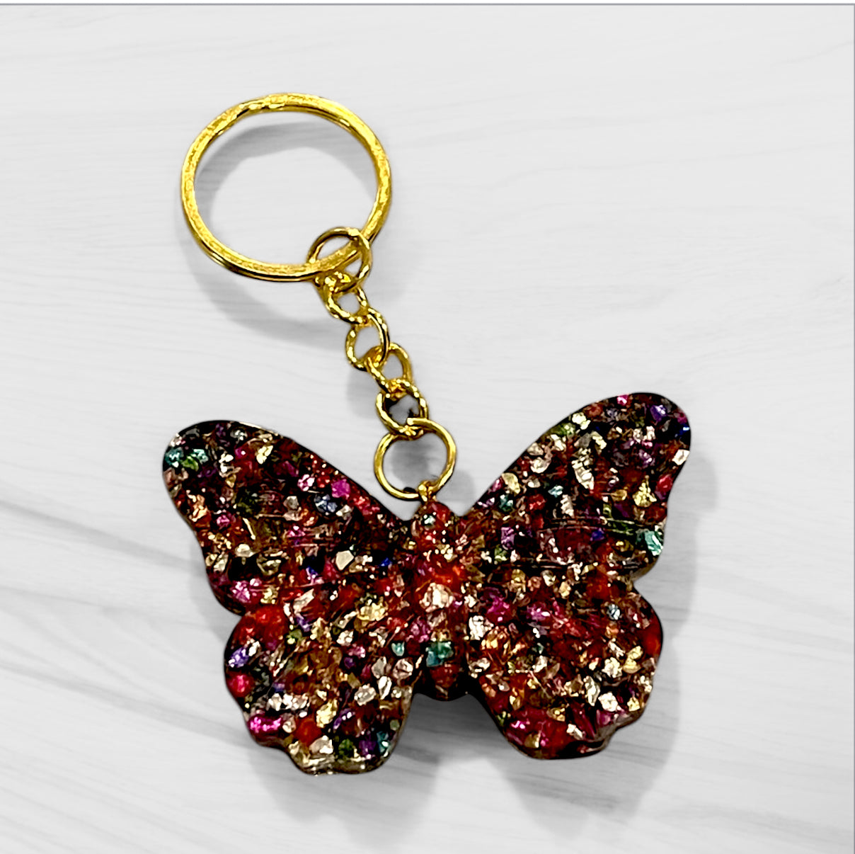 Handmade butterfly resin keychain with multi-colored metallic flakes in red, gold, and rainbow tones, featuring gold-tone hardware and chain detail